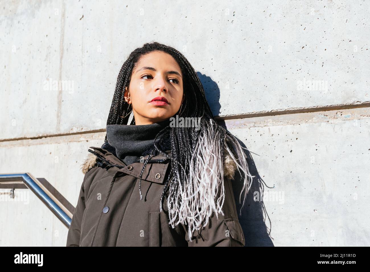 Paisible femme avec de longs cheveux tressés et dans des vêtements chauds se tenant près du mur dans la rue et regardant loin Banque D'Images