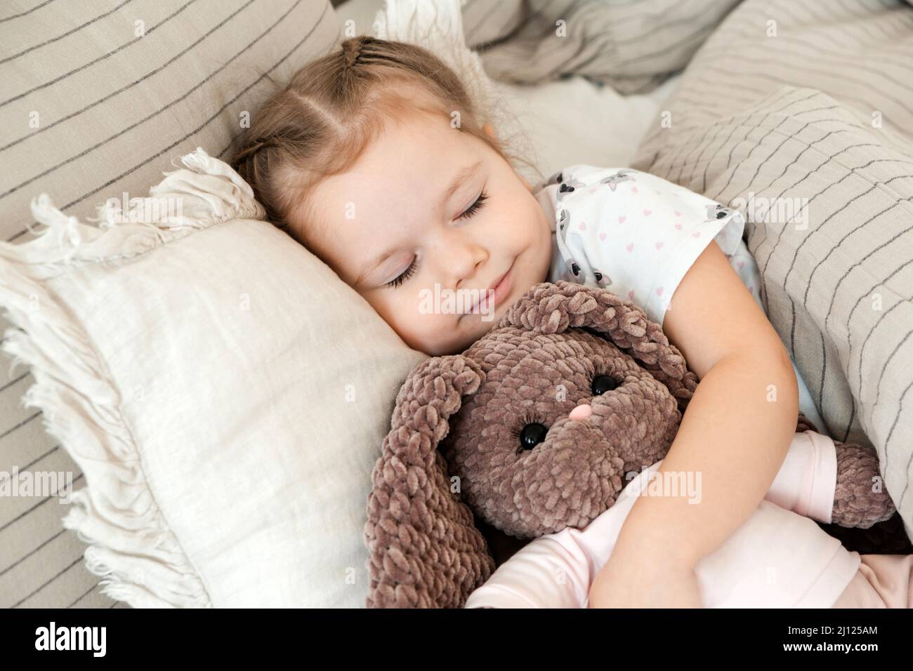 Belle petite fille embrassant un lapin en peluche tout en dormant dans le lit à la maison. Sommeil paisible de bébé. Banque D'Images