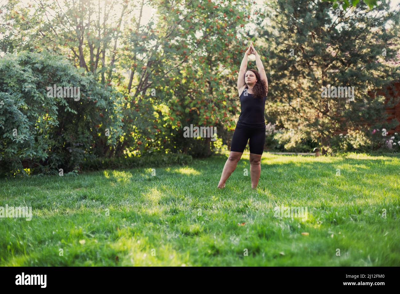 Femme surpondérée tenant les mains en s'étirant en faisant du yoga en appréciant le moment sur l'herbe verte sur l'arrière-cour du cottage avec maison en bois et arbres dedans Banque D'Images