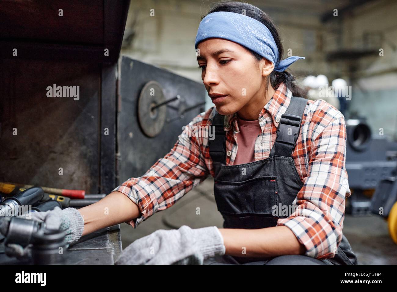 Jeune femme contemporaine en vêtements de travail en choisissant des pièces de rechange métalliques tout en allant réparer une machine industrielle dans un grand atelier Banque D'Images