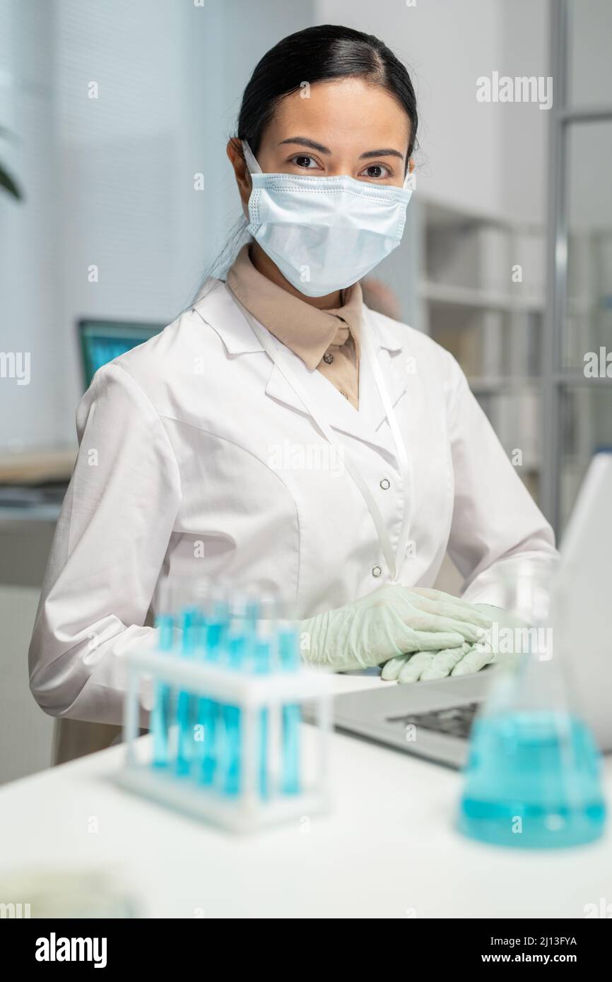 Jeune scientifique féminine en whitecoat, gants et masque de protection assis près d'un bureau devant un ordinateur portable et jeu de flacons avec du liquide bleu Banque D'Images