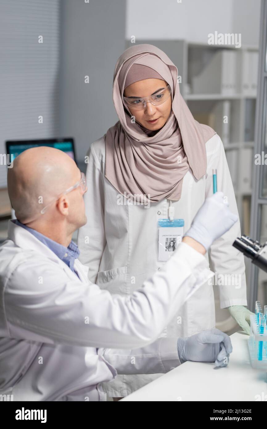 Jeune virologue musulmane de hijab et de witecavoine regardant le liquide bleu dans la fiole tenue par un collègue mâle mature en laboratoire Banque D'Images