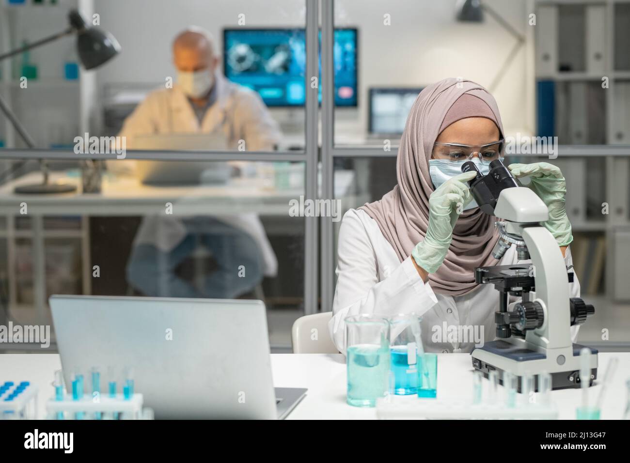 Jeune virologiste musulmane féminine étudiant les caractéristiques du nouveau virus au microscope contre la mise en réseau de collègues mâles en laboratoire Banque D'Images