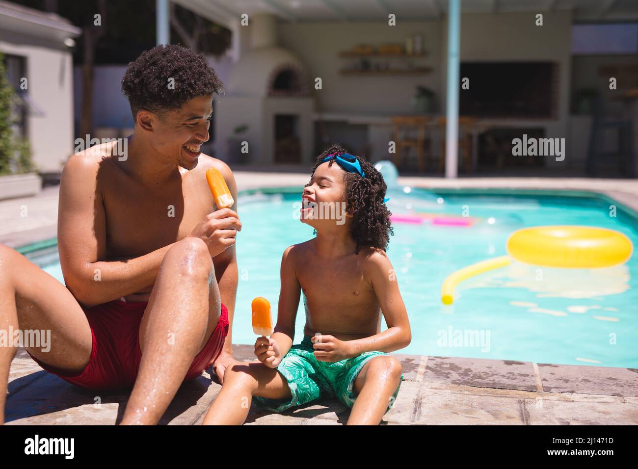 Joyeux père et fils hispaniques mangeant des glaces tout en étant assis au bord de la piscine par beau temps Banque D'Images