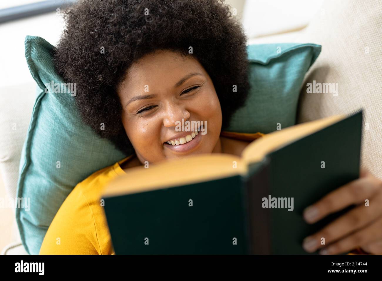 Gros plan à grand angle souriant afro-américain femme adulte de taille moyenne lisant un livre tout en étant couché sur un canapé Banque D'Images