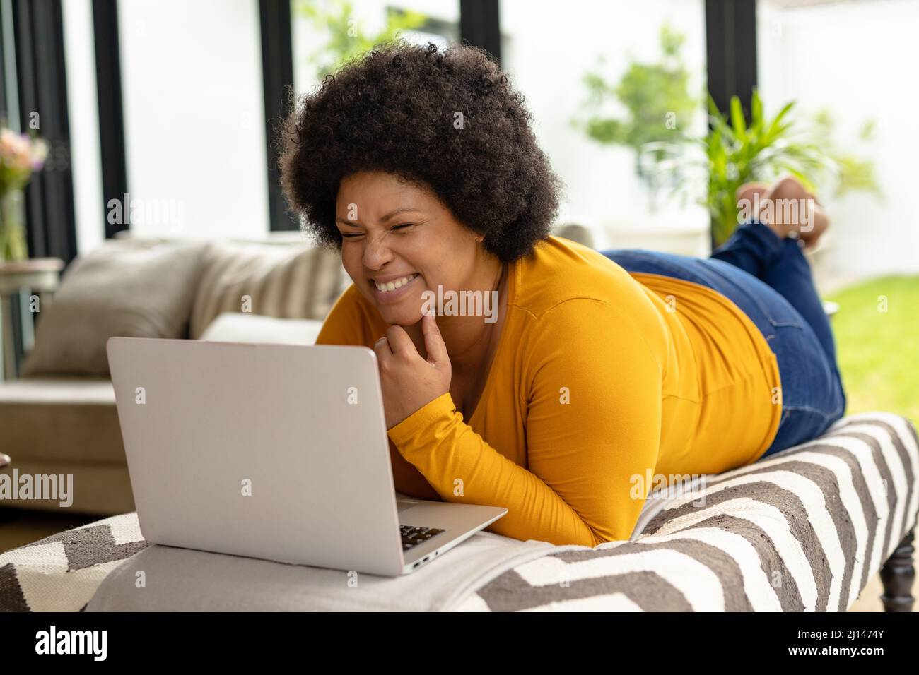 Femme afro-américaine souriante utilisant un ordinateur portable tout en étant allongé sur un canapé à la maison Banque D'Images