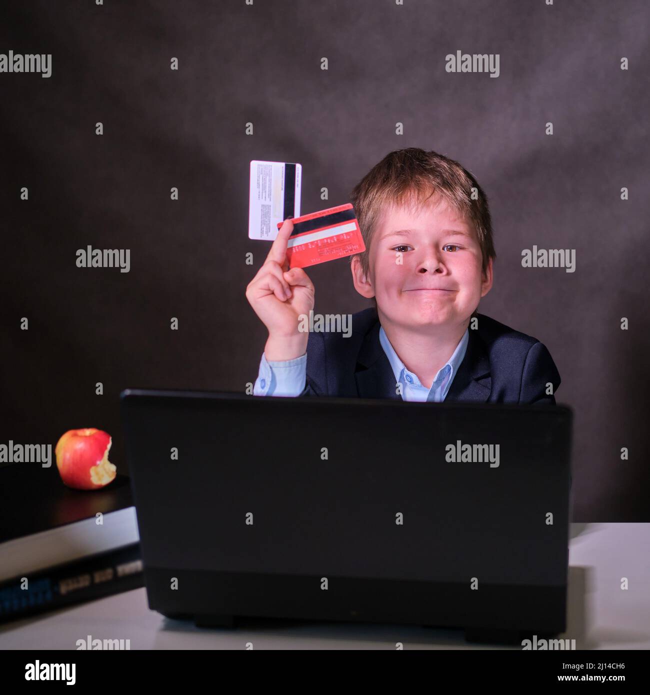 Un garçon souriant dans un costume d'école avec des cartes de crédit à l'ordinateur, un espace de copie sur un arrière-plan sombre de studio Banque D'Images