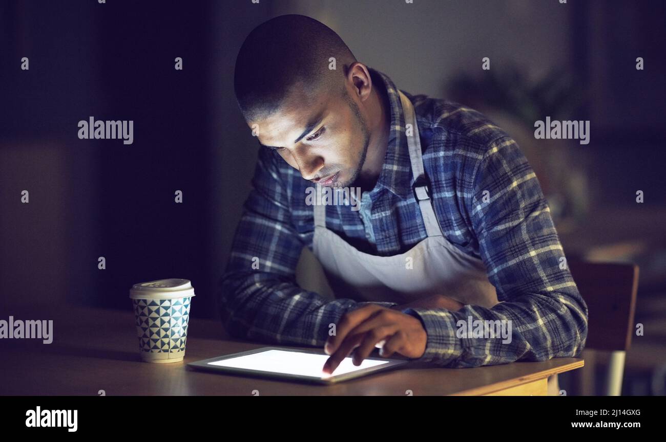 Il a l'entrainement de faire des tâches à tout moment. Photo d'un jeune homme travaillant tard sur une tablette numérique dans son café. Banque D'Images