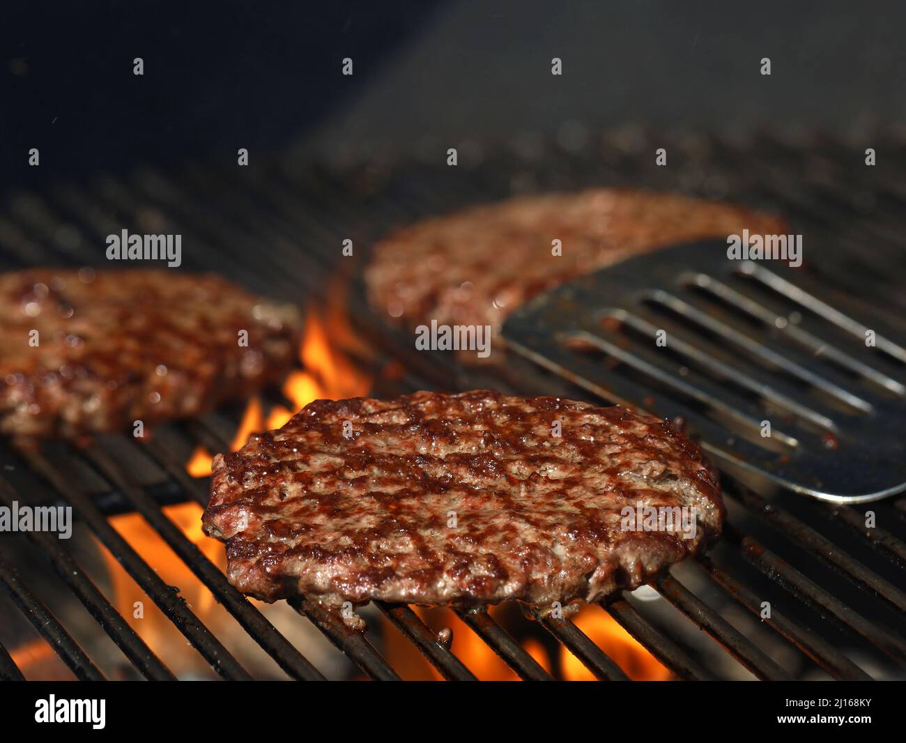 Hamburgers de bœuf fraîchement grillés sur grille avec flammes et spatule en acier inoxydable en arrière-plan Banque D'Images