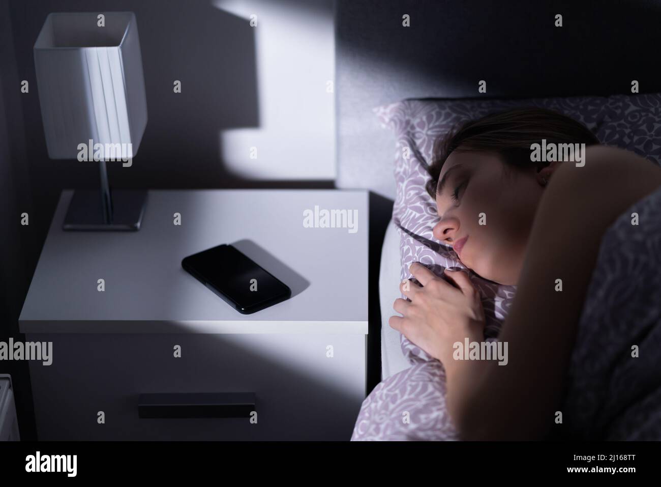Femme dormant dans le lit avec le téléphone sur la table la nuit. Téléphone portable sur la table de nuit. Smartphone avec mode silencieux, silencieux ou éteint à côté de la personne au repos. Banque D'Images
