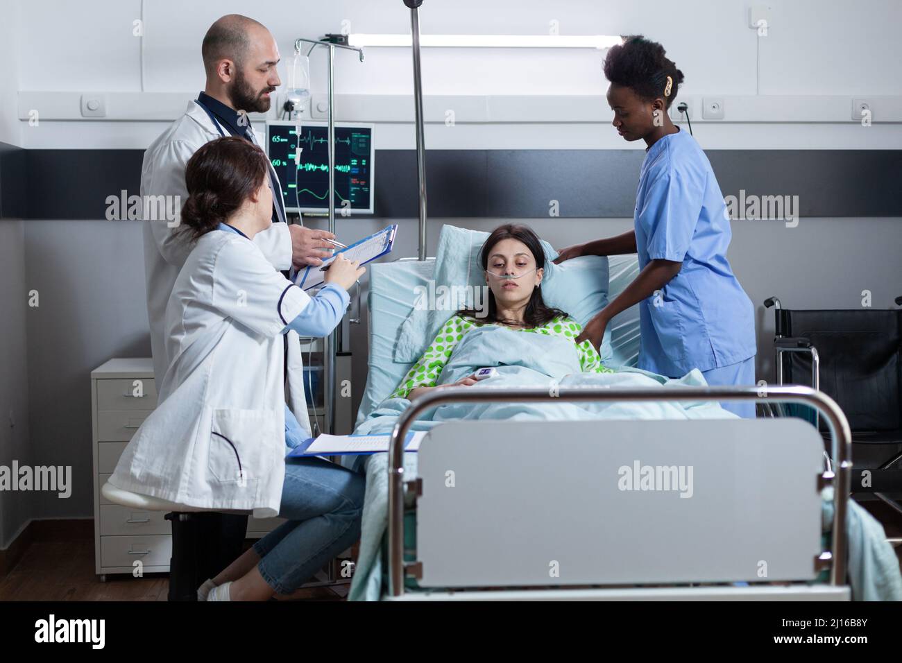 Équipe de médecins discutant des antécédents médicaux du patient sur le presse-papiers avant la chirurgie pendant que l'infirmière donne des soins médicaux. Femme avec une faible saturation de SpO2 connectée à une ligne d'égouttage iv et un moniteur affichant les paramètres vitaux. Banque D'Images