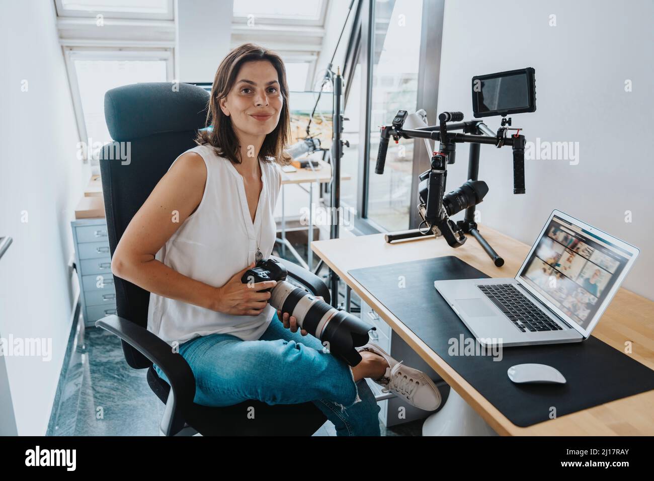 Photographe souriant avec appareil photo numérique assis sur une chaise et ordinateur portable sur une table au studio Banque D'Images
