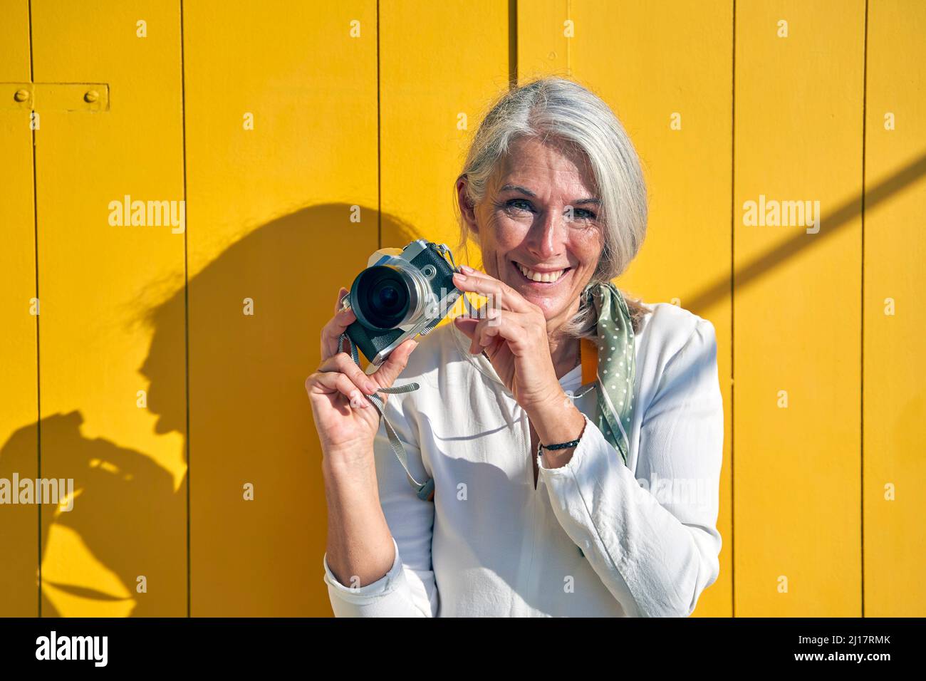 Femme souriante montrant un appareil photo debout devant la porte jaune par beau temps Banque D'Images