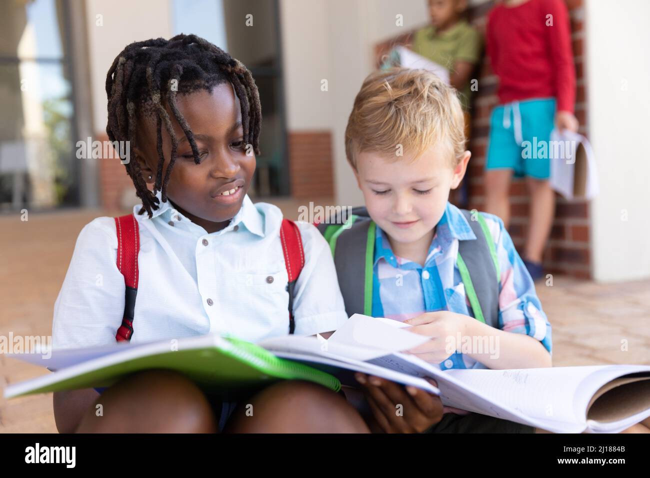 Élèves multiraciaux de l'élémentaire étudiant à partir de livres tout en étant assis sur le sol à l'entrée de l'école Banque D'Images