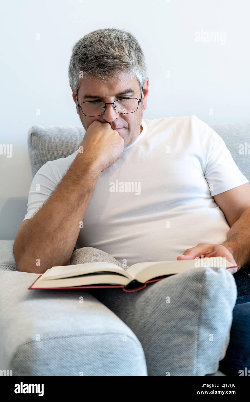 Homme aux cheveux gris avec des lunettes assis dans un fauteuil lisant un livre. Concept de détente à la maison Banque D'Images