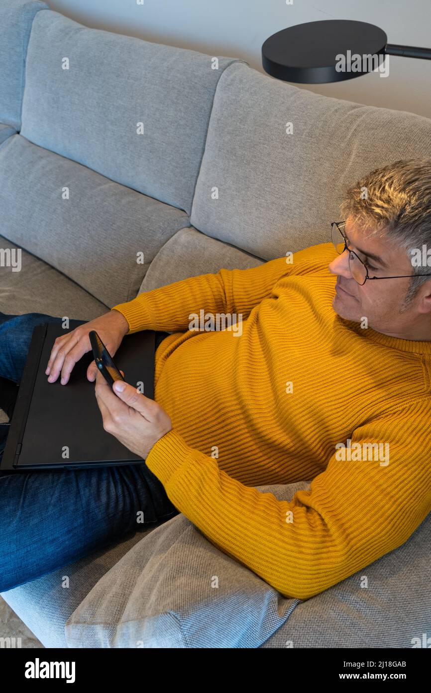 Homme assis sur un canapé à la maison SMS sur un téléphone avec ordinateur portable fermé. Repos à la maison Banque D'Images