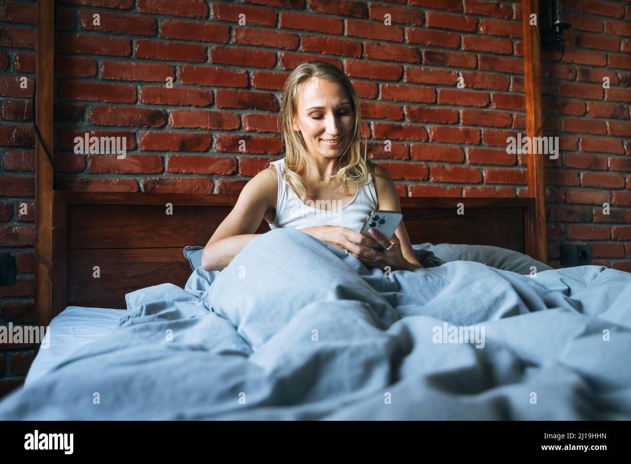 Femme adulte de quarante ans avec cheveux longs blonds en pyjama utilisant le téléphone portable assis sur le lit dans la chambre loft à la maison Banque D'Images