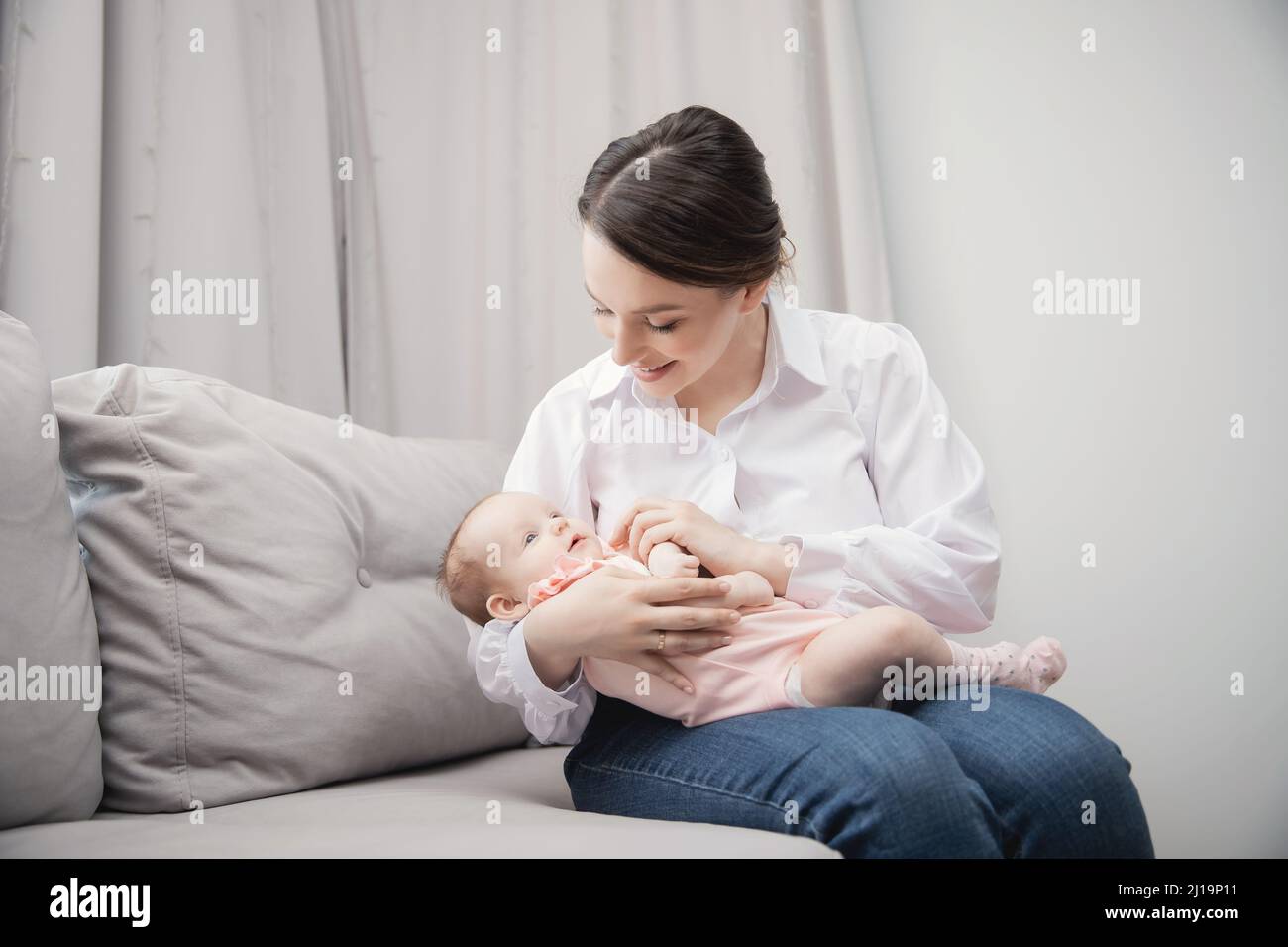 Bonne jeune mère femme tenant bébé fille dans ses bras, se préparant à l'allaitement. Intérieur de l'appartement avec canapé gris. Banque D'Images