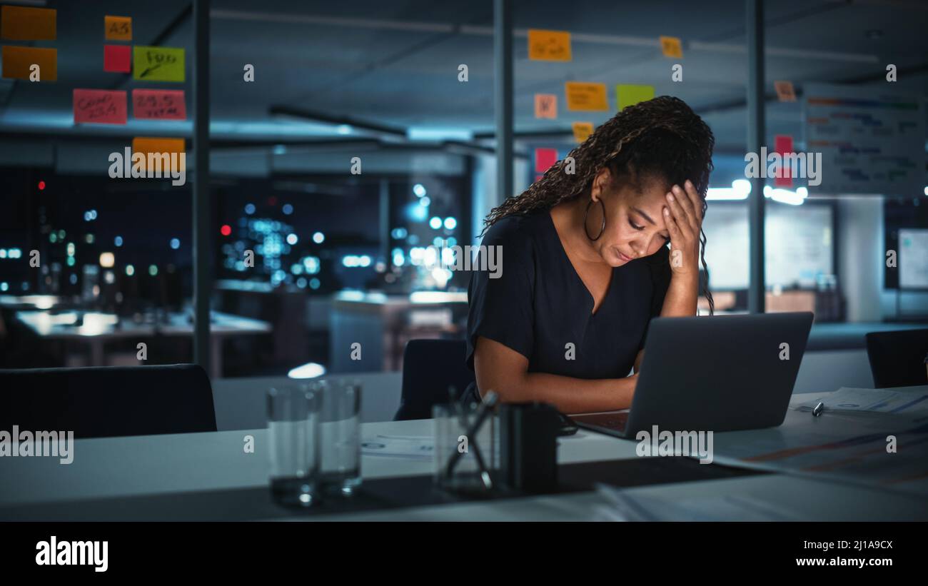 Une femme d'affaires afro-américaine surtravaillée travaille sur un ordinateur portable au bureau de Big City tard dans la soirée. Fatigué stressé femme Entrepreneur essayer Banque D'Images