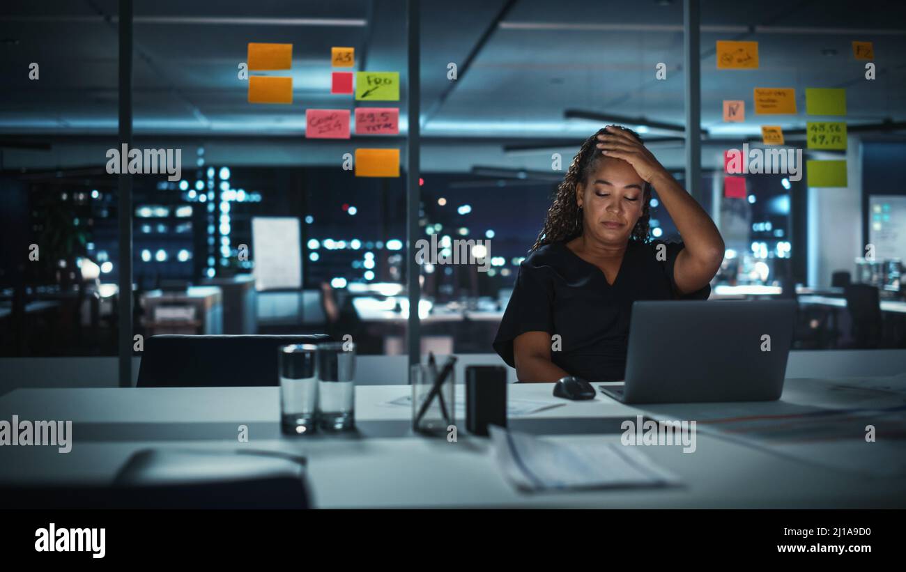 Une femme d'affaires afro-américaine surtravaillée travaille sur un ordinateur portable au bureau de Big City tard dans la soirée. Fatigué stressé femme Entrepreneur essayer Banque D'Images