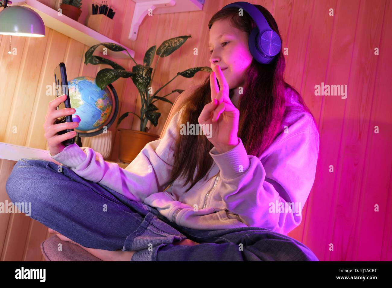 Joyeux mignon joli adolescent dans un casque moderne joyeux, dans la pièce au néon à la maison. Jeune fille qui fait du selfie, écoutez votre musique préférée, passez un appel vidéo. Amuse-toi bien Banque D'Images