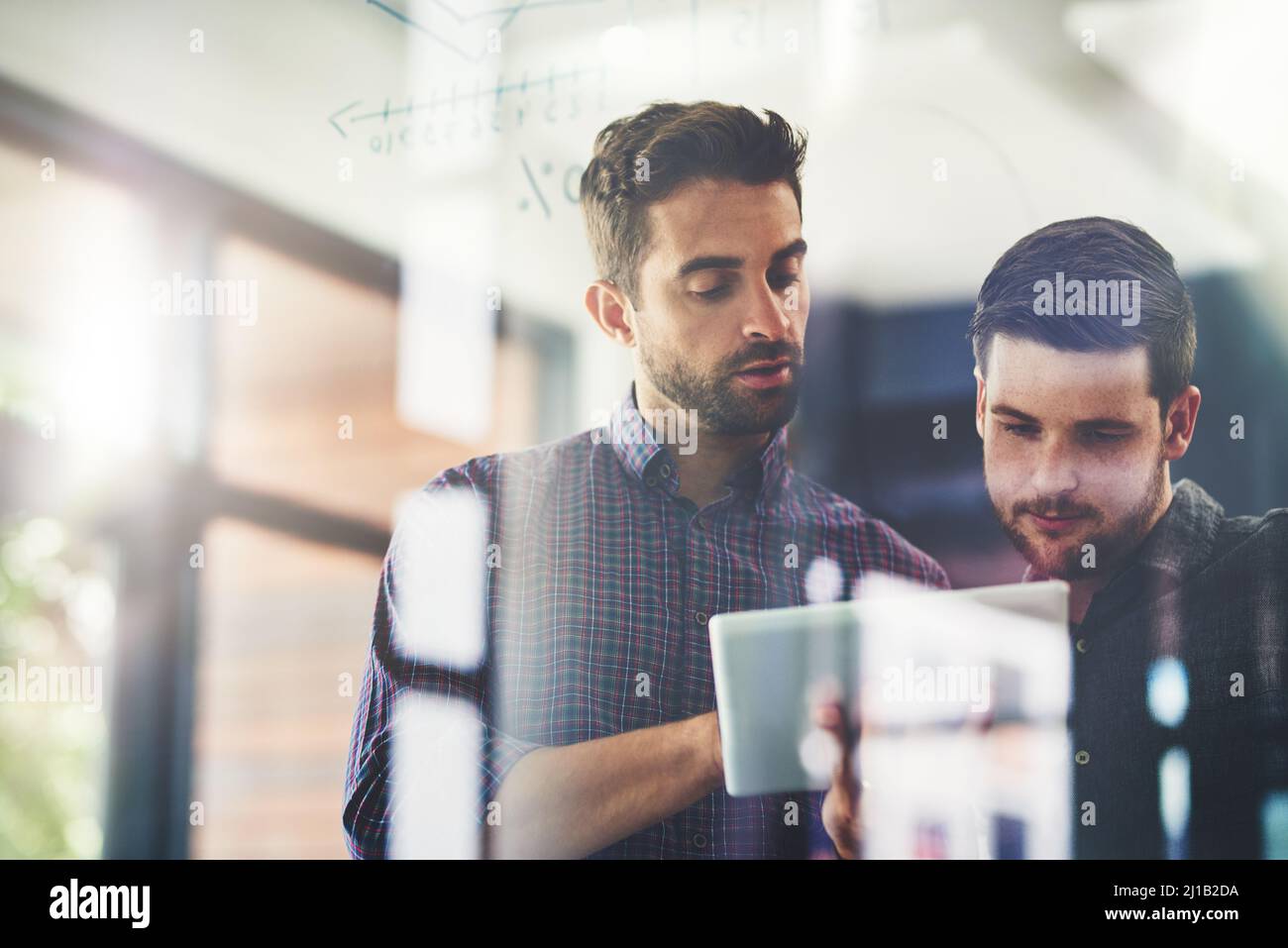 Travailler sur leurs idées principales pour développer leur entreprise. Tourné de deux jeunes hommes d'affaires utilisant une tablette numérique tout en brainstorming sur un mur de verre dans un Banque D'Images