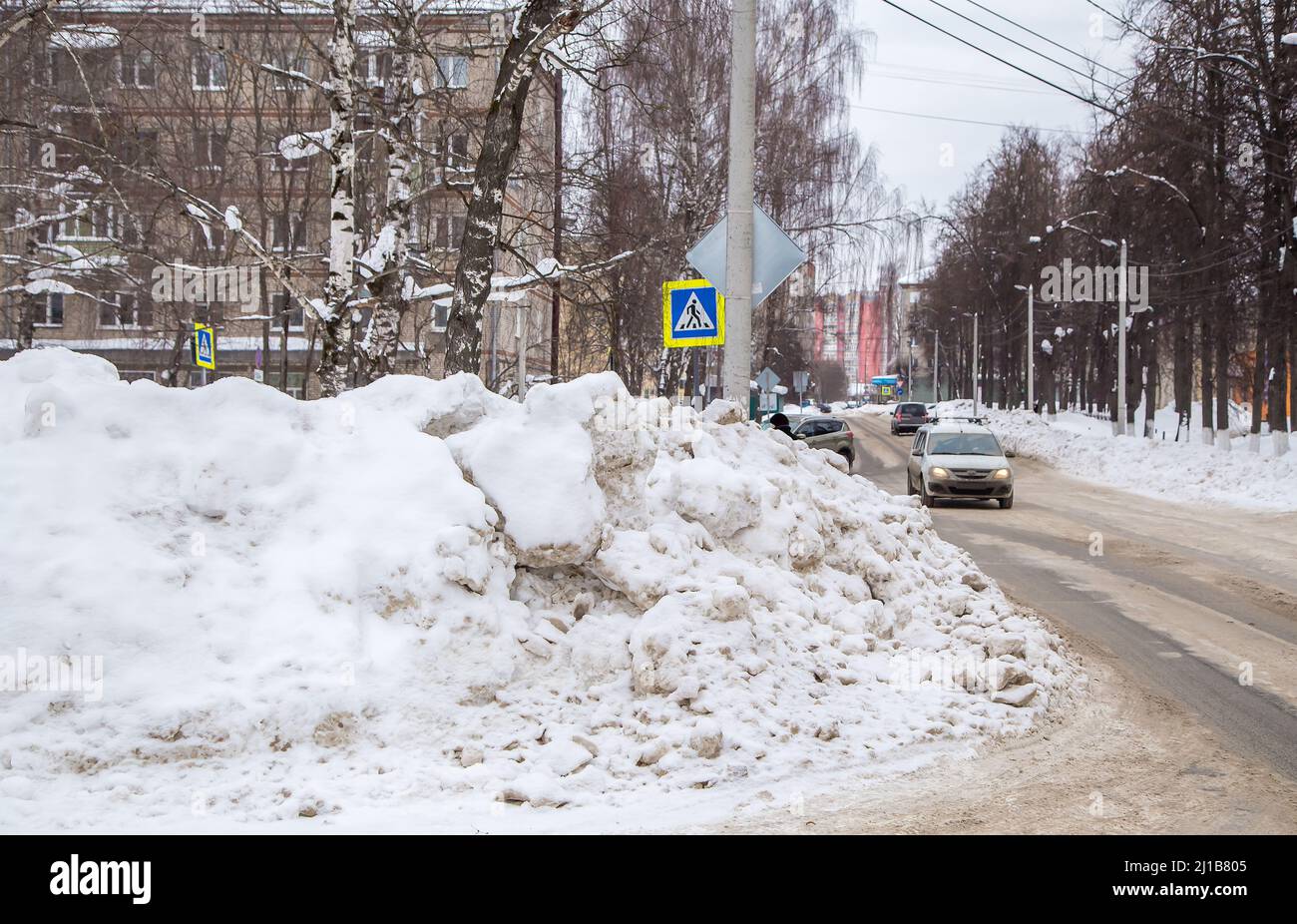 Une énorme dérive de neige au bord de la route sur fond de rue de la ville. Sur la route se trouve de la neige sale dans des tas hauts. Paysage urbain d'hiver. Jour d'hiver nuageux, lumière douce. Banque D'Images