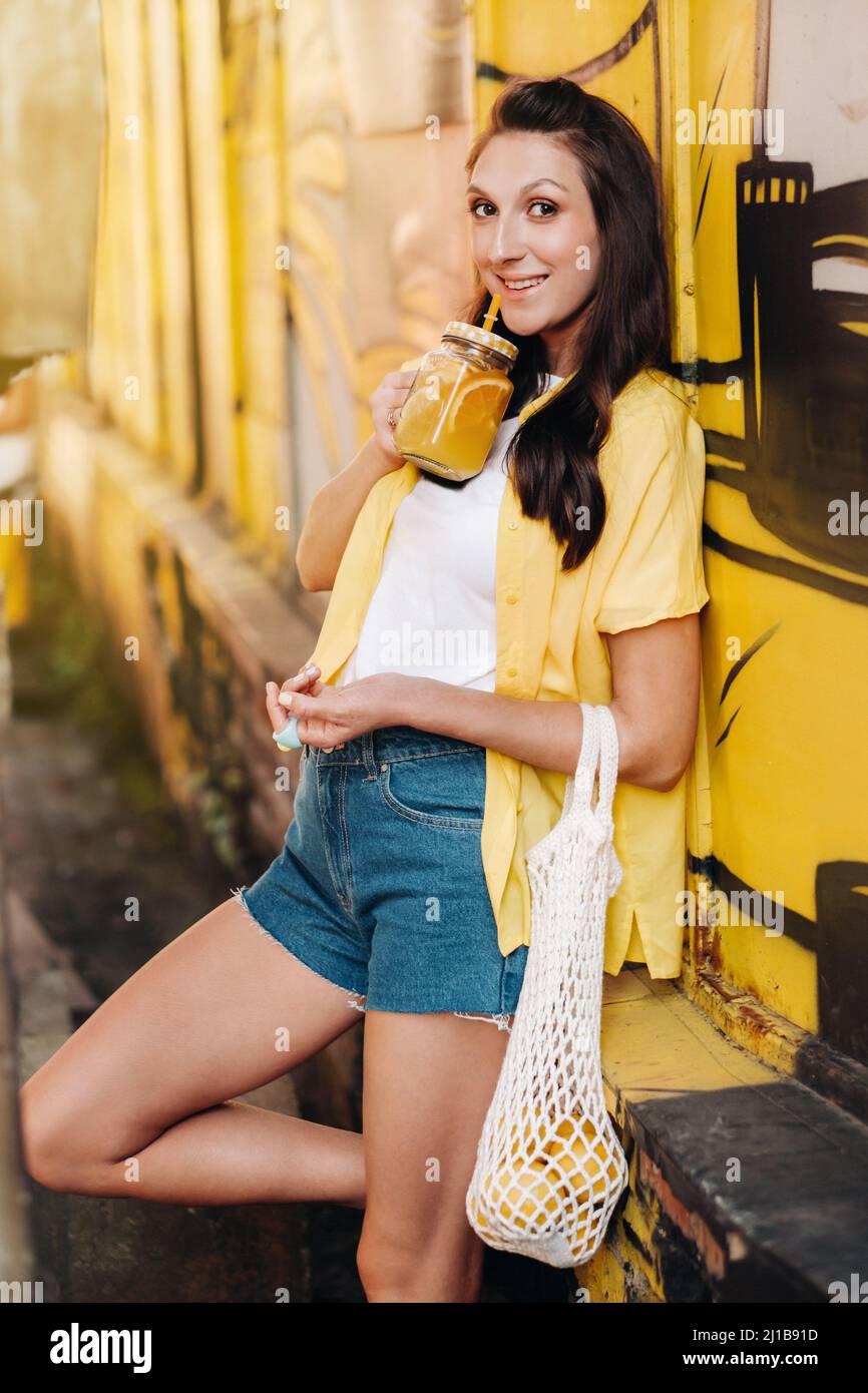 une fille dans une chemise jaune, un short et des chaussures noires avec un  filet de citrons et jus de citron marche le long d'une rue de ville. Une  femme avec un