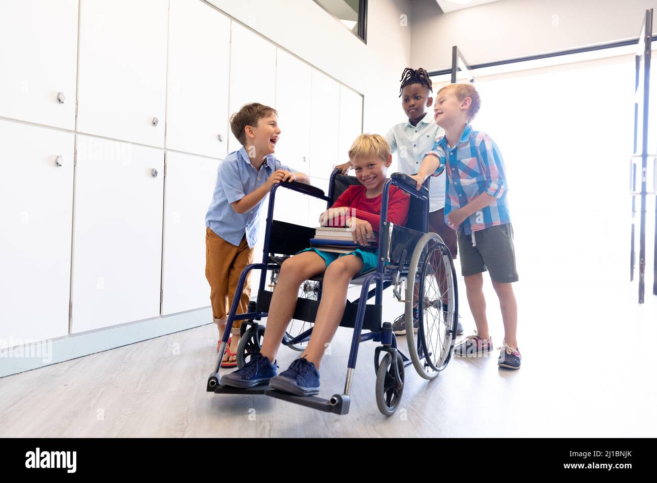 Des écoliers gais et multiraciaux debout par un camarade de classe masculin assis sur un fauteuil roulant dans le couloir de l'école Banque D'Images