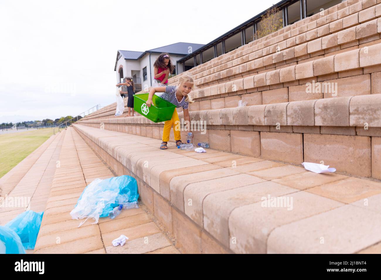 Les écolières élémentaires multiraciales nettoient les déchets plastiques sur les marches de construction d'écoles contre le ciel Banque D'Images