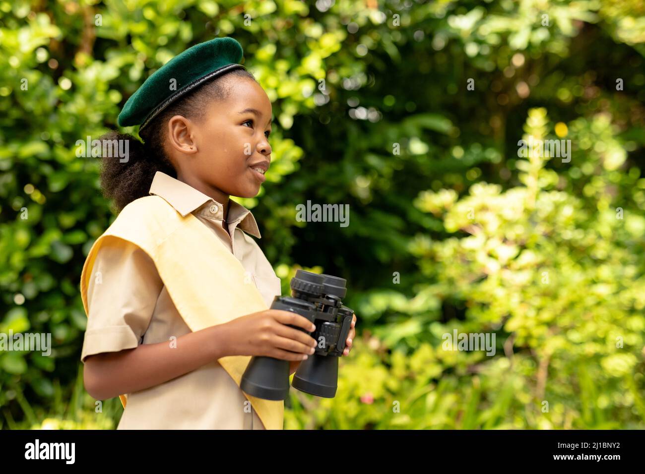 Souriante fille scout afro-américaine en uniforme tenant des jumelles dans la forêt Banque D'Images