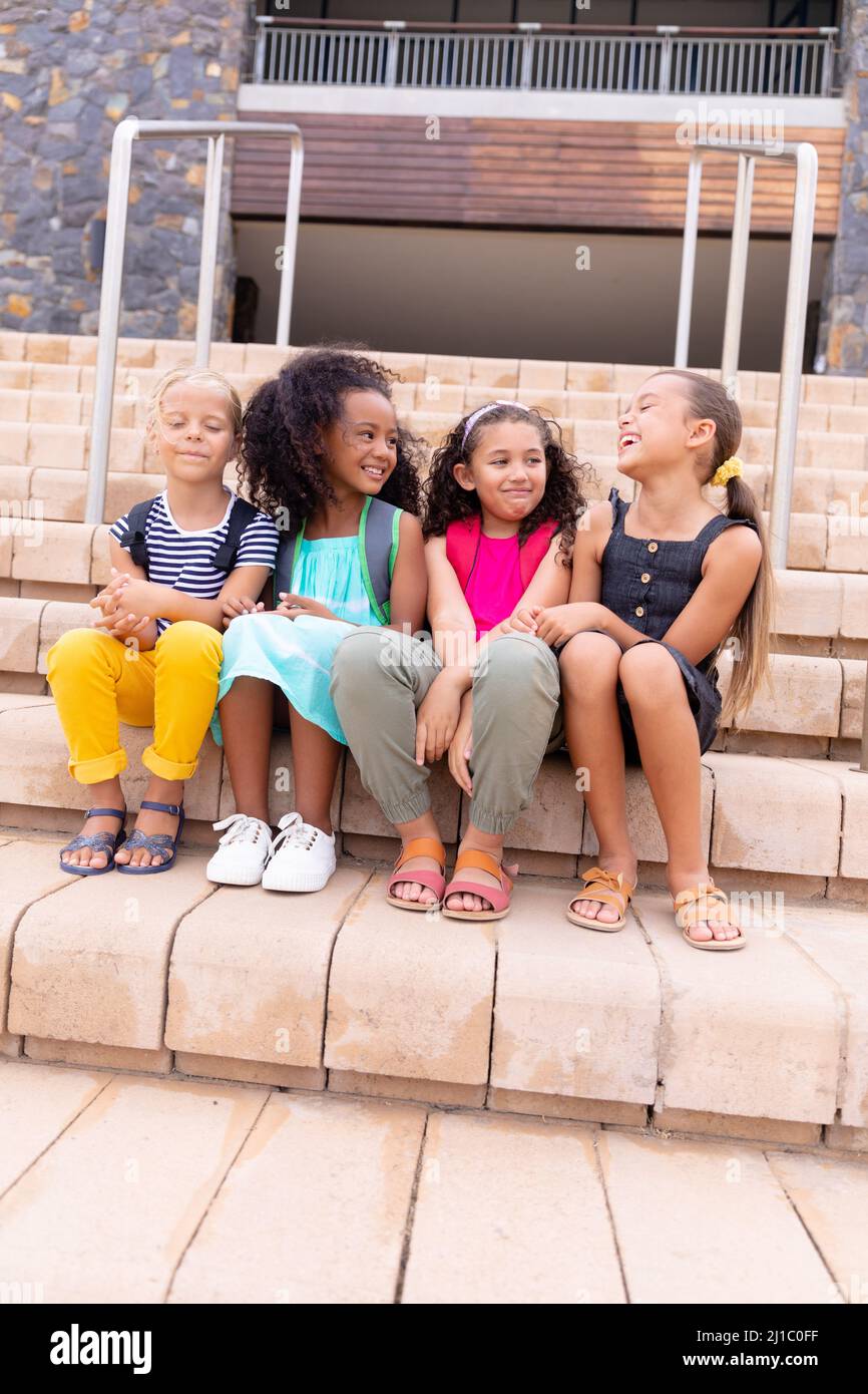 Une longueur complète de jeunes filles élémentaires souriantes et multiraciales assises sur des marches contre le bâtiment de l'école Banque D'Images