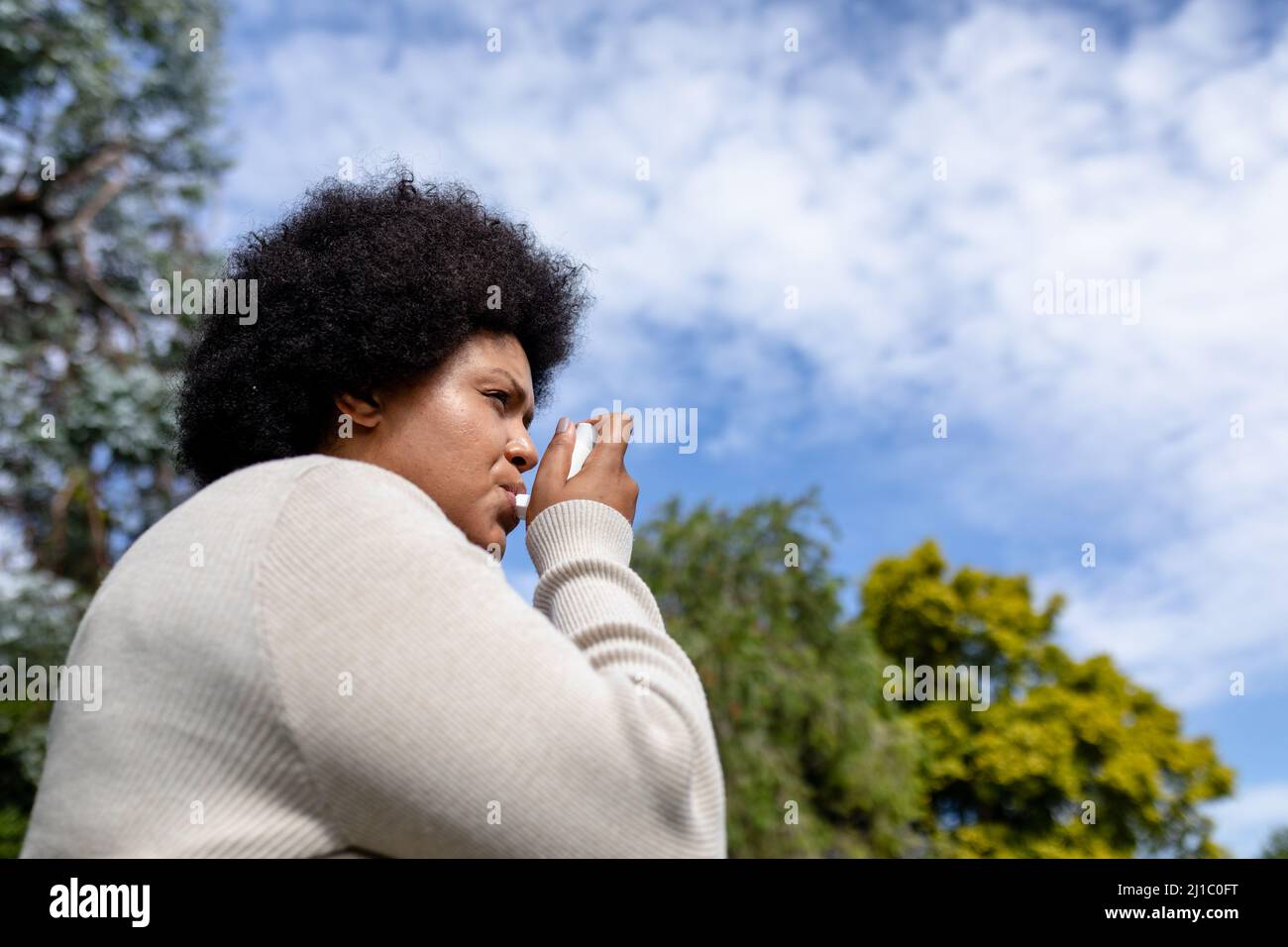 Vue à angle bas de la femme afro-américaine moyenne adulte utilisant un inhalateur d'asthme tout en se tenant contre le ciel Banque D'Images