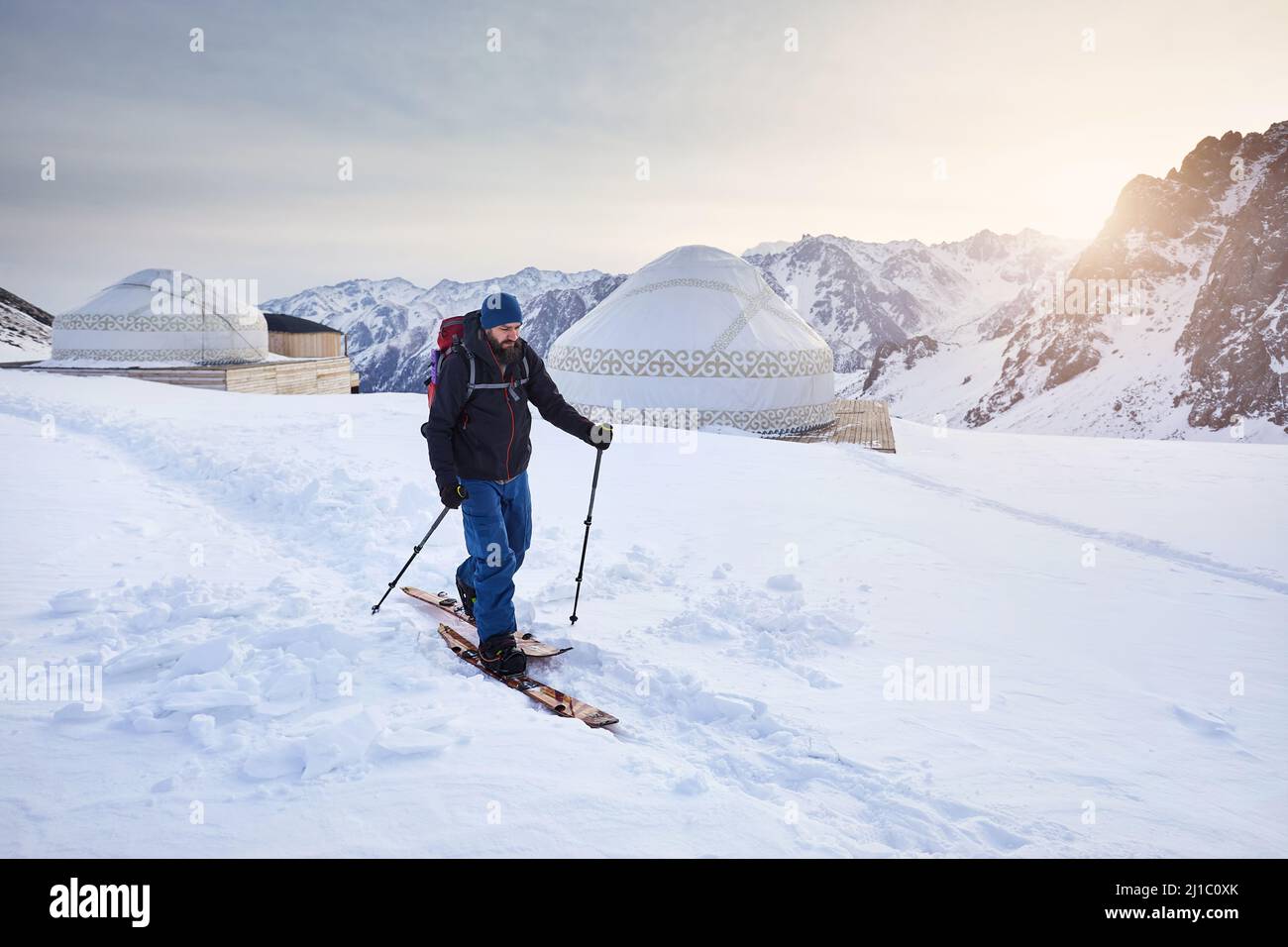 Skieur homme ski en montant avec la barbe et le sac à dos à haute montagne enneigée près de la maison de yourte traditionnelle nomade à la station de ski de Shymbulak à Almaty, Kazak Banque D'Images