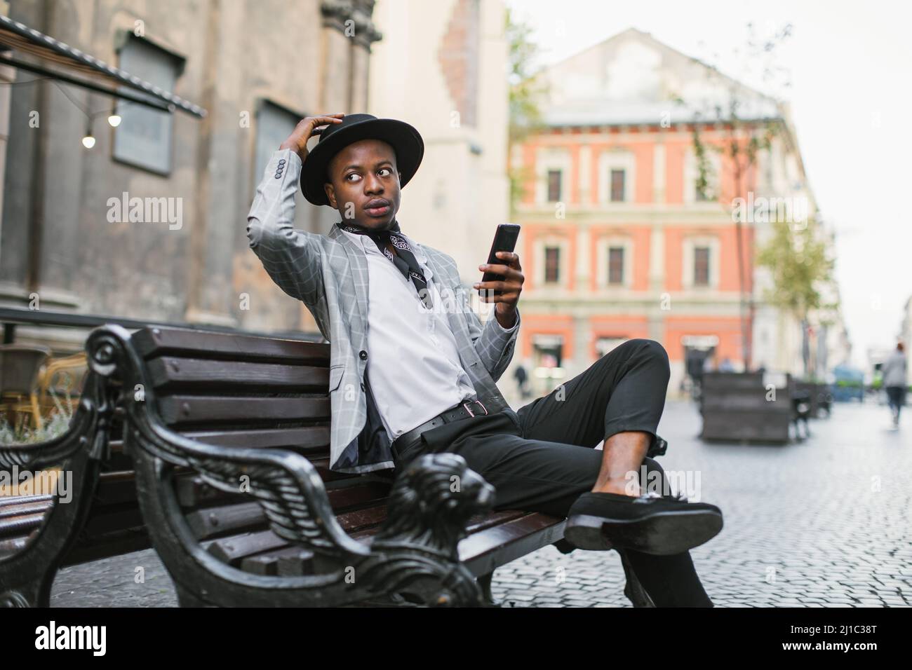 Homme afro-américain en costume élégant et chapeau regardant de côté en  étant assis sur un banc avec un smartphone moderne dans les mains. Concept  de technologie et de personnel Photo Stock -