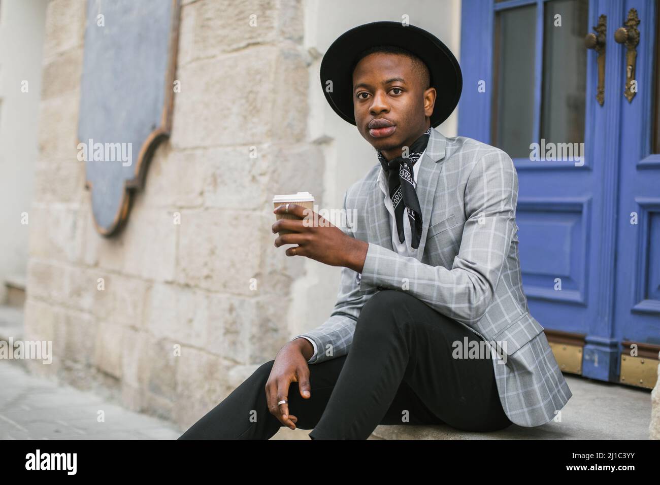 Beau homme africain portant un costume élégant et un chapeau noir assis à  l'extérieur avec une tasse de café. Jeune homme d'affaires prospère  regardant la caméra avec confiance dans les yeux Photo