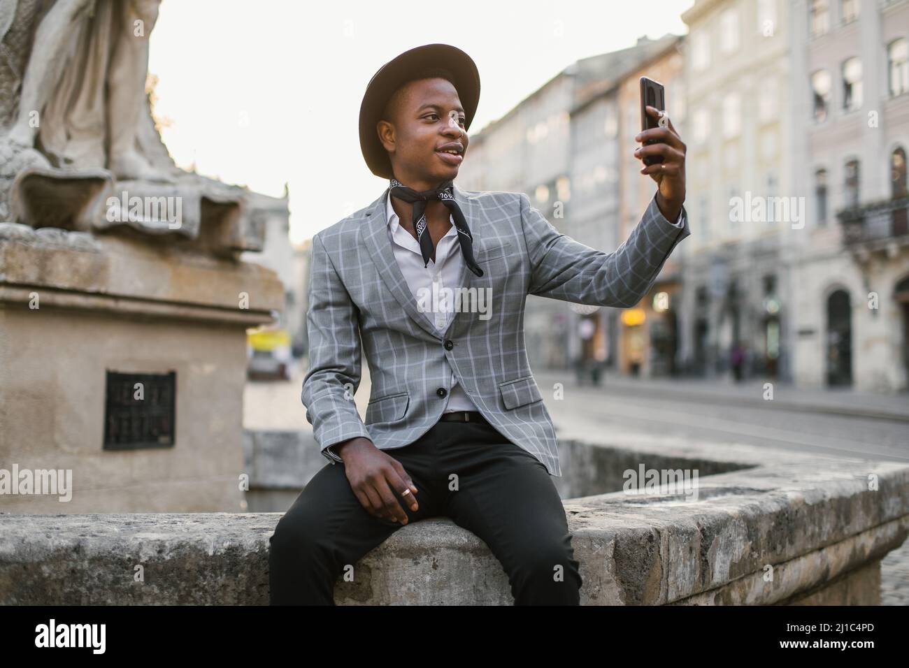 Un homme afro-américain portant un costume et un chapeau élégants utilisant  un smartphone moderne pour emporter son selfie tout en se reposant à  l'extérieur. Concept des personnes, de la technologie et des