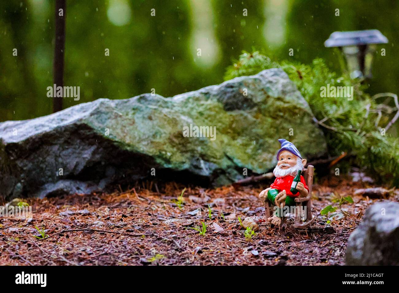 Un gnome de pelouse reposant sur un banc au milieu de la pluie entouré de rochers. Banque D'Images