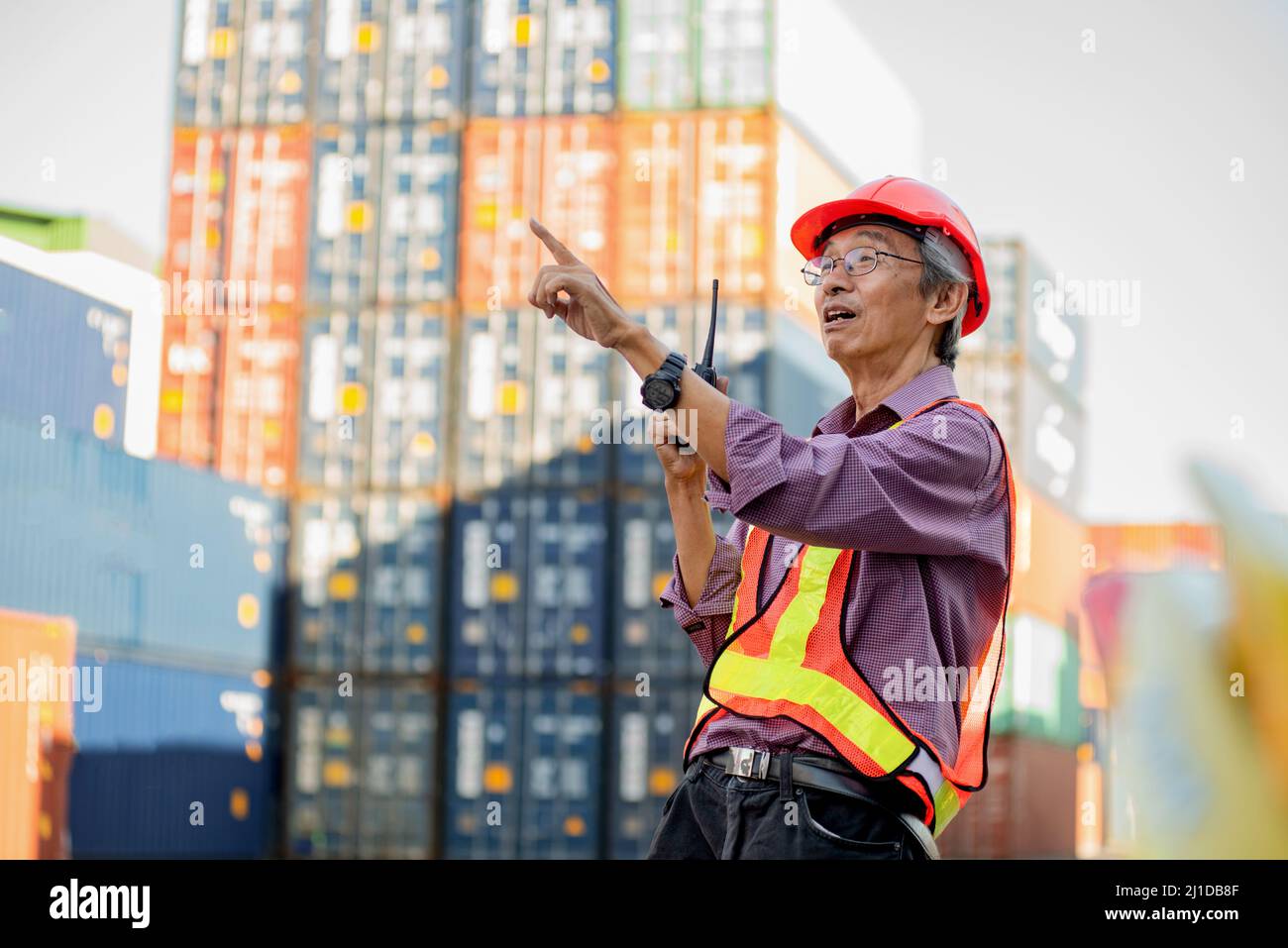 Un ingénieur travailleur asiatique âgé de longue date portant un gilet de sécurité et un casque debout et tenant des radios-walkies talkie à des conteneurs de fret d'expédition logistique Banque D'Images