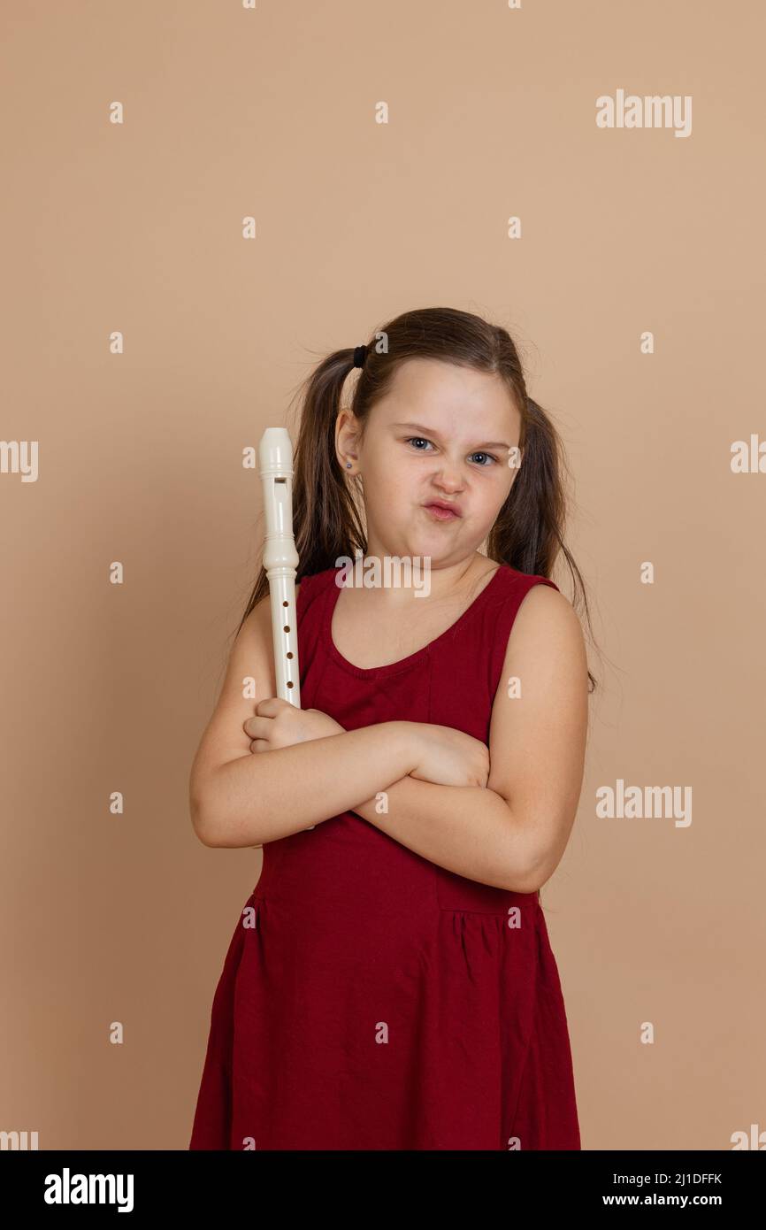 Fille en robe rouge avec l'expression mécontente tenir flûte avec les bras croisés, fond beige. Apprendre à jouer de l'instrument de musique de vent de bois. Banque D'Images