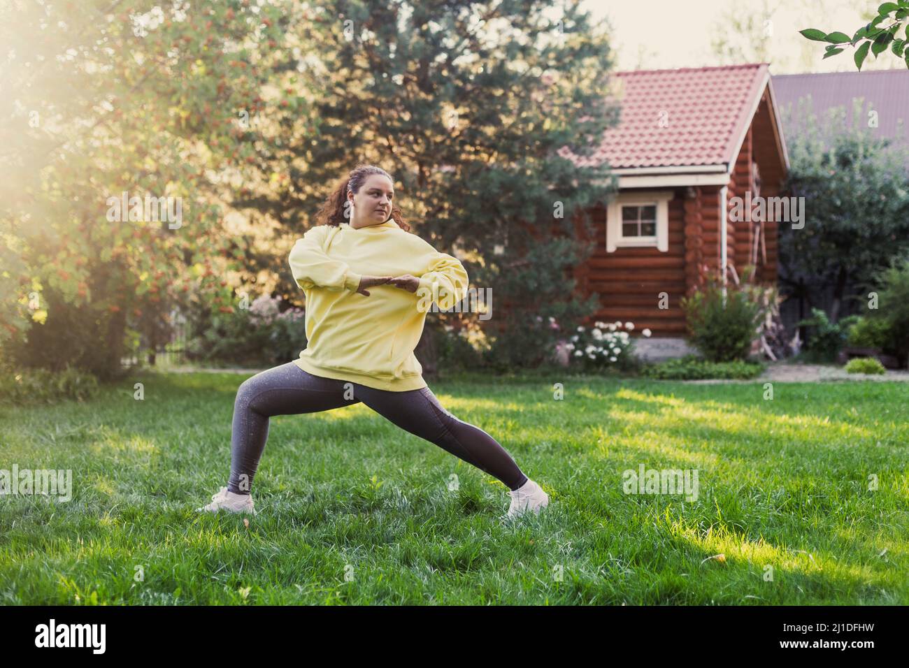 Femme d'âge moyen adulte faisant des exercices physiques s'étirant pour garder la forme en portant des vêtements de sport sur l'arrière-cour sur l'herbe verte avec maison de campagne en bois Banque D'Images