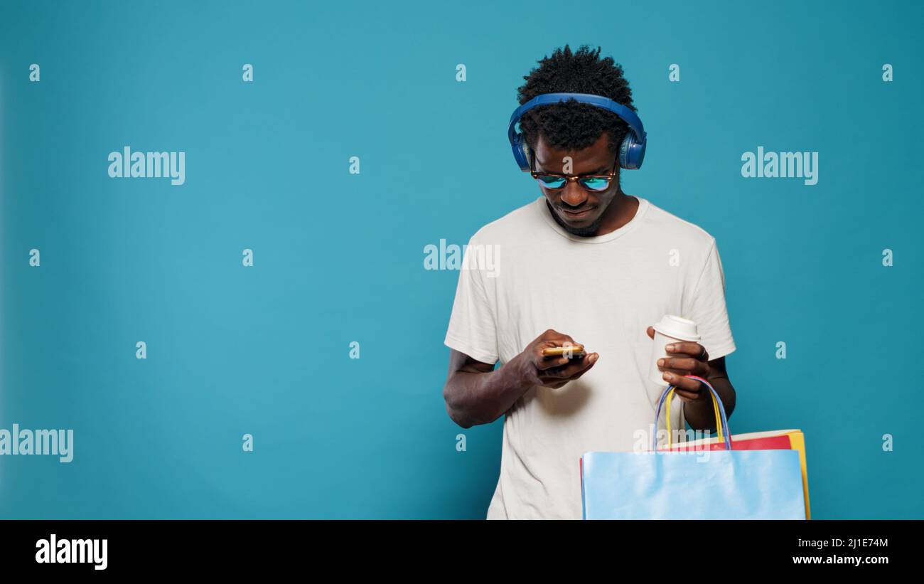 Adulte moderne utilisant un smartphone et transportant des sacs de shopping dans un magasin. Jeune homme avec des lunettes de soleil écoutant de la musique sur des écouteurs et regardant le téléphone après l'achat de vêtements Banque D'Images