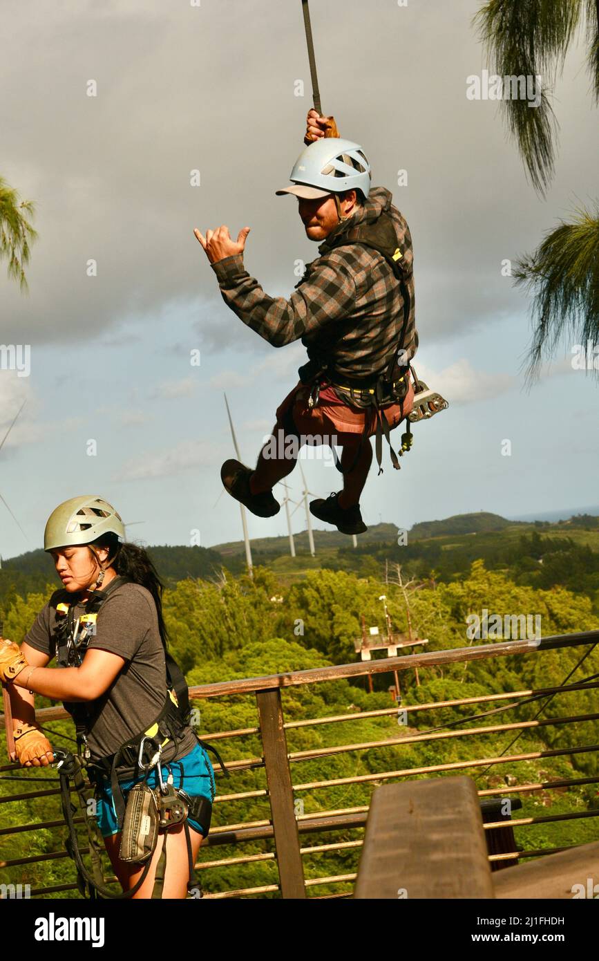 Homme donnant le geste de shaka tout en se repoussant vers le bas de la plate-forme, dans le cadre d'une excursion aventure en tyrolienne à Climbworks Keana Farms, North Shore, Kahuku, HI, États-Unis Banque D'Images