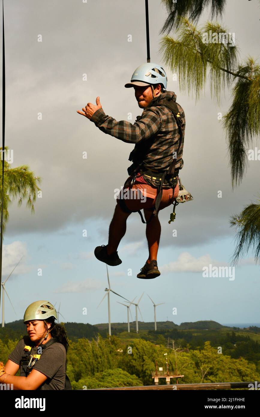 Homme donnant le geste de shaka tout en se repoussant vers le bas de la plate-forme, dans le cadre d'une excursion aventure en tyrolienne à Climbworks Keana Farms, North Shore, Kahuku, HI, États-Unis Banque D'Images