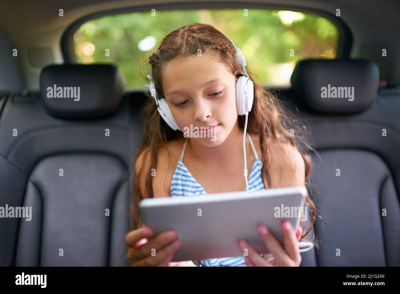 Regarder ses films sur la banquette arrière. Photo d'une jeune fille assise dans un siège arrière de voiture avec un casque et une tablette numérique. Banque D'Images