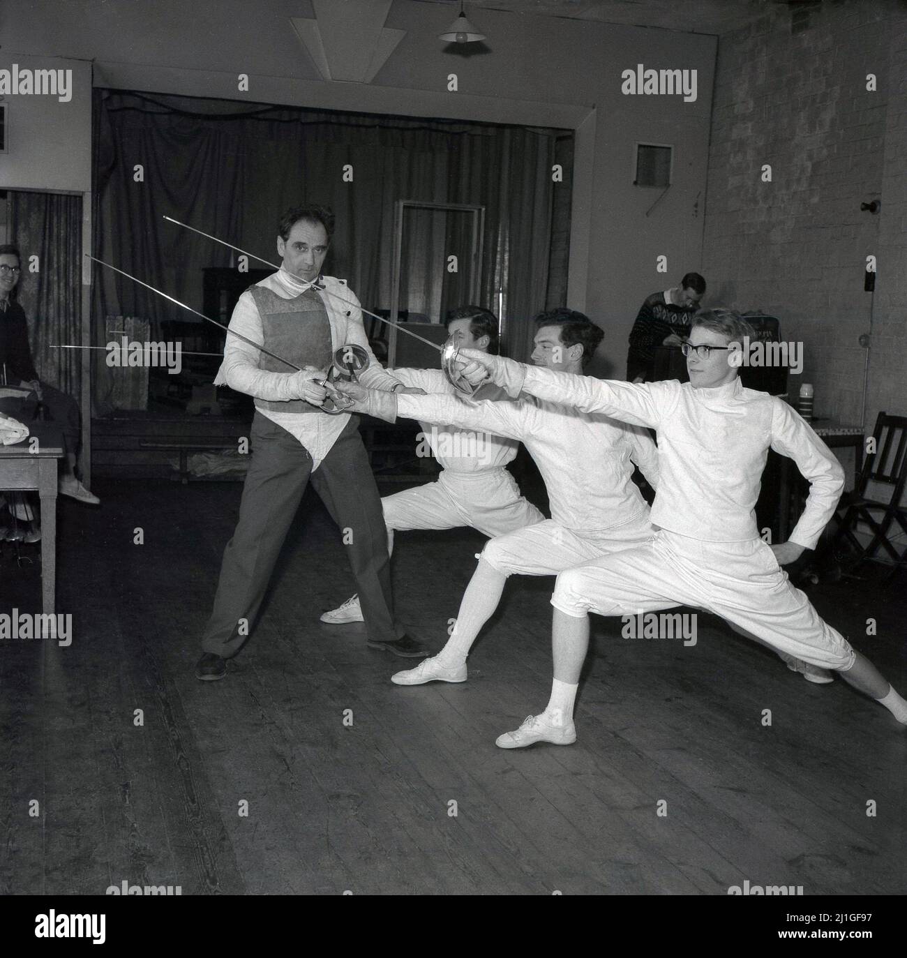 1960s, étudiant historique, masculin portant des culottes, des vestes et des pilmolos traditionnels dans une classe d'escrime, avec instructeur, Université d'Oxford, Angleterre, Royaume-Uni. Ils travaillent sur la poussée des tireurs, un mouvement clé dans le sport, qui est une poussée d'attaque faite avec un pied en avant et la jambe arrière droite et le bras d'épée tendu vers l'avant. Banque D'Images