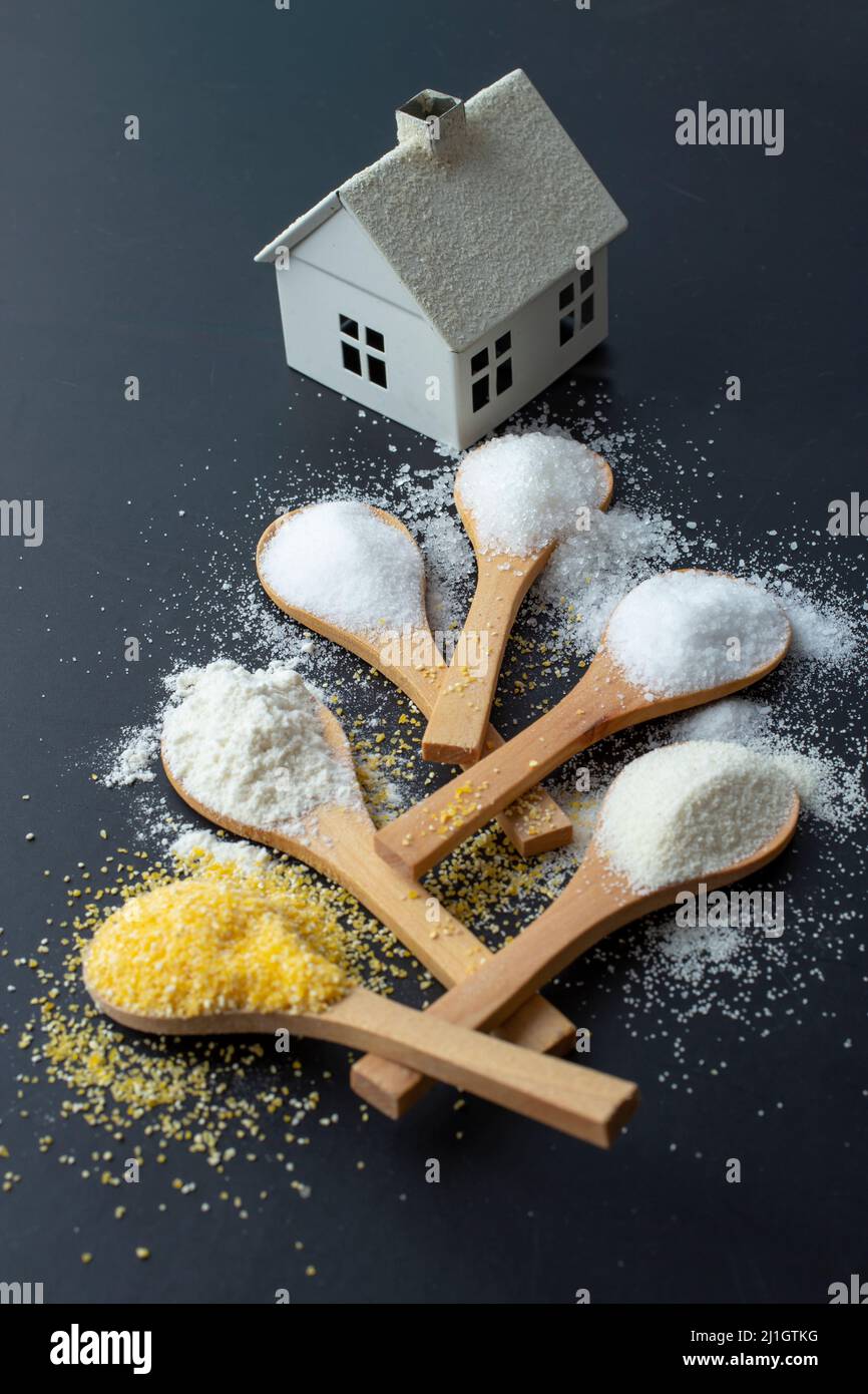 Cuillères à café en bois avec farine, sel, sucre, semoule et farine de maïs, menant à une maison blanche miniature Banque D'Images