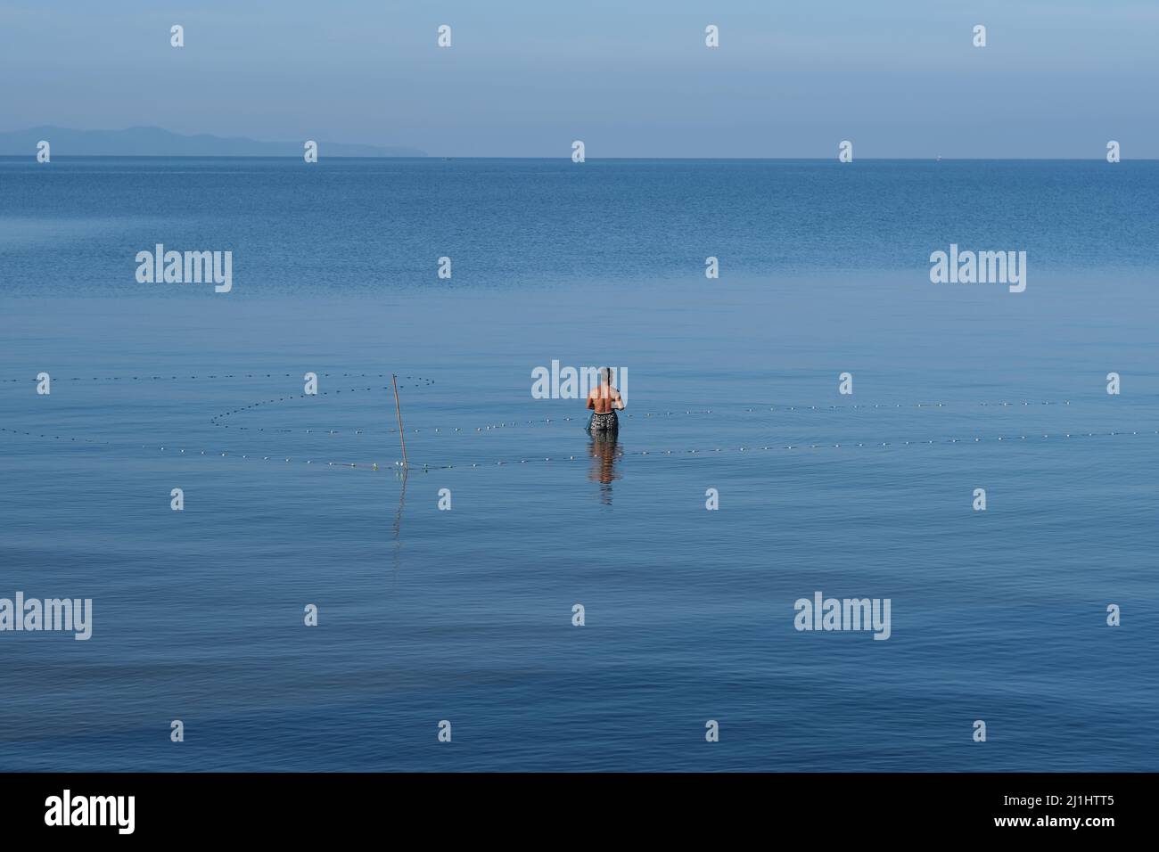 Un pêcheur solitaire avec filet de pêche se dresse au milieu d'une mer et d'un ciel bleu sans limites Banque D'Images
