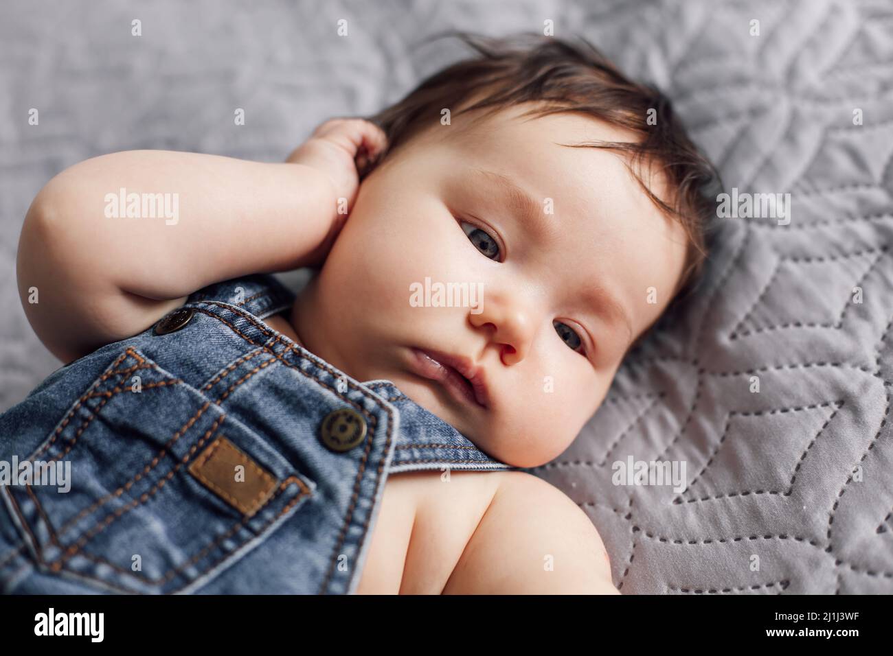 Portrait de bébé portant une barboteuse en denim reposant sur une couverture grise à la maison. Un bébé calme s'est couché dans la chambre, explorez le monde. Gris Banque D'Images