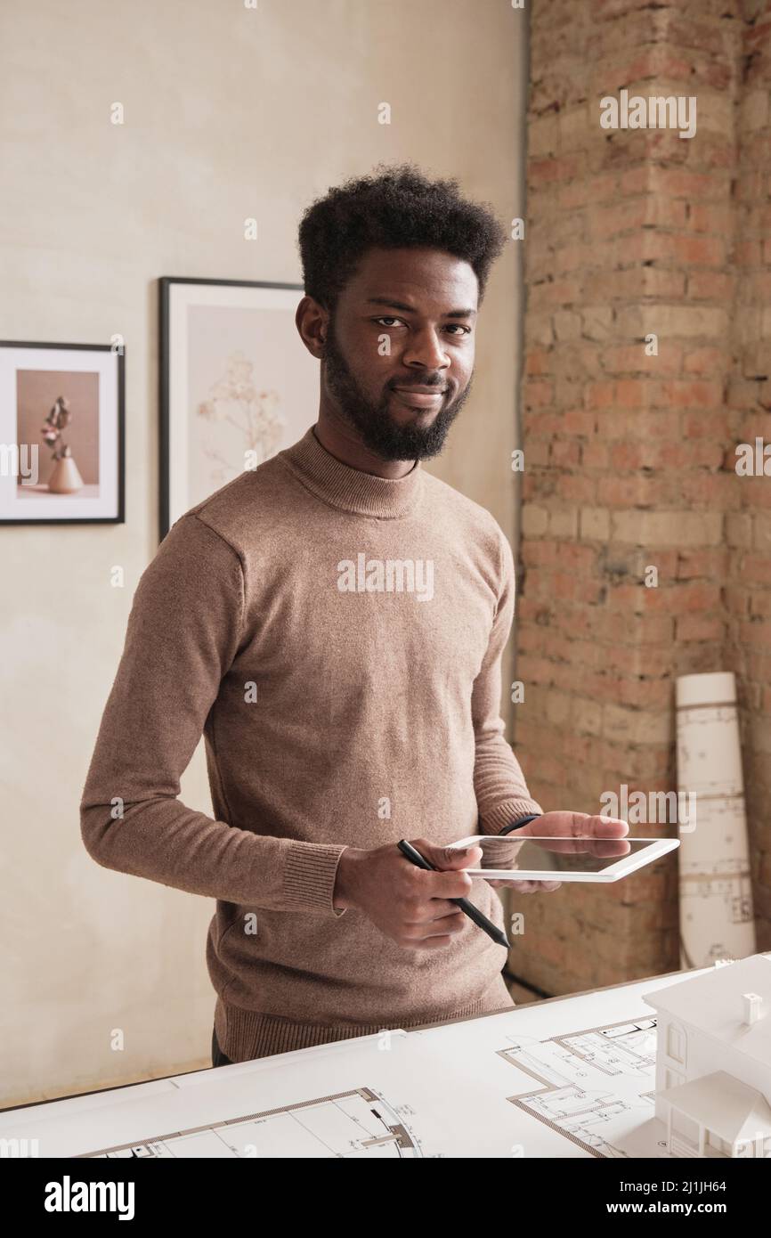 Portrait d'un jeune architecte noir souriant avec une barbe debout à table avec plan et à l'aide d'une tablette tout en élaborant la conception du bâtiment Banque D'Images